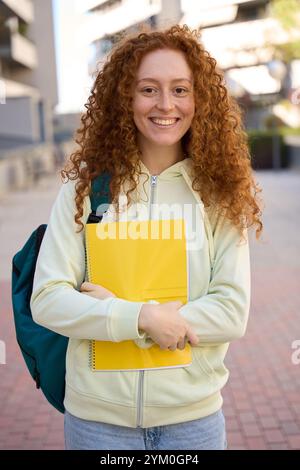 Smiling student holding a yellow notebook and wearing backpack in university campus Stock Photo