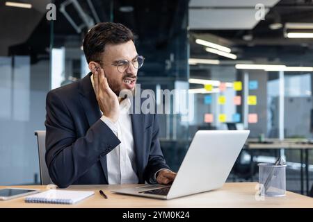 Businessman with beard in office feeling ear pain during online meeting. Wearing glasses and suit, sitting at desk with laptop and notepad on wooden table. Stock Photo