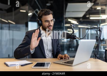 Man in office records podcast using microphone and laptop, wearing headphones. Engages audience through camera, embodying modern work culture, communication, and technology trends. Stock Photo