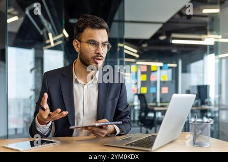 Young professional in modern office discusses business strategies using laptop and notebook. Engaging in thoughtful communication in collaborative work environment. Stock Photo