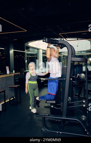 Senior sportive man in sportswear training in gym, doing pull-up exercises in modern gym with special equipment. Elderly woman waiting her turn Stock Photo
