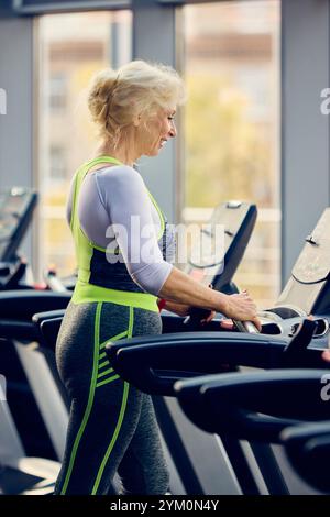 Senior woman with blonde hair, in comfortable sportswear training, exercising on treadmill in brightly lit gym. Taking care after health and body Stock Photo