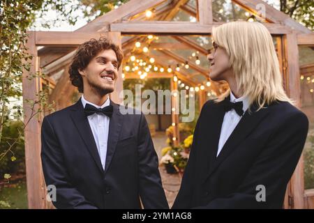 Two young men in formal attire share a joyful moment in a beautifully lit outdoor setting. The enchanting garden is adorned with twinkling lights, cre Stock Photo