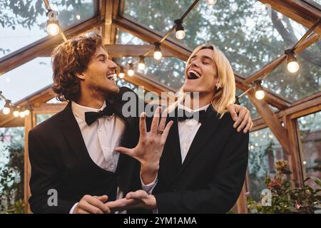 Two young men share a joyful moment in a beautifully decorated greenhouse, surrounded by plants and warm lights. Their laughter resonates, celebrating Stock Photo