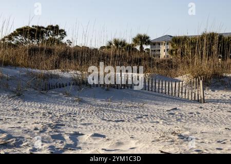 A wooden fence is set along the sand dunes, surrounded by tall grass and a beachside building nearby on Hilton Head Island in South Carolina. Stock Photo