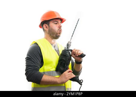 Adult builder man blowing air on drill to cool down wearing orange safety helmet and fluorescent vest as work equipment isolated on white studio backg Stock Photo