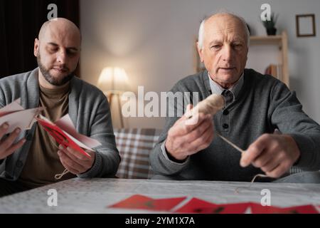 Senior Father and young adult son prepare decorations for the holiday, crafting garlands and decor, enjoying family fun sitting in the living room at Stock Photo