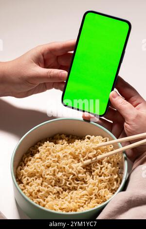 Female using smartphone while eating pasta at home. Smartphone with green screen with space for text. Smartphone in hands with served table in backgro Stock Photo