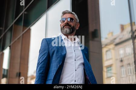 A handsome stylish mature man wearing sunglasses and suit while walking in the city street near the urban building. Stock Photo