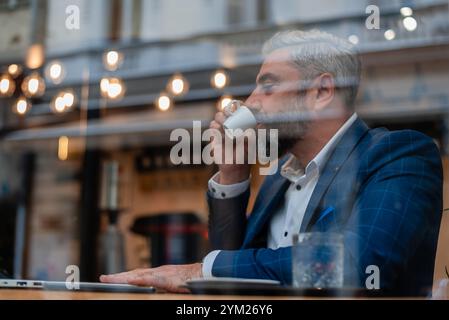 A handsome mature business man sitting at his desk in the office or restaurant cafe near the window drinking a coffee and working on a laptop. Stock Photo