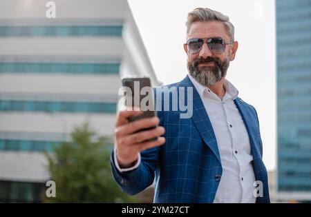 A happy mature business man using a cell phone app texting outside of the office in the city with urban buildings in the background. Stock Photo