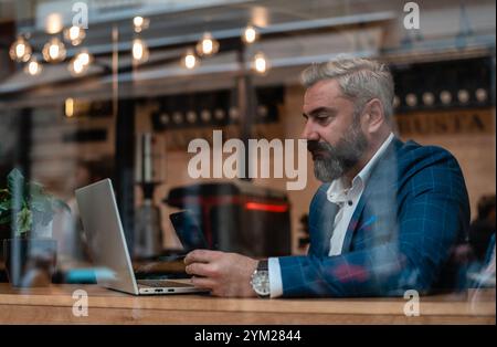 A handsome mature business man using his phone while working on a laptop in the office near the window. Stock Photo