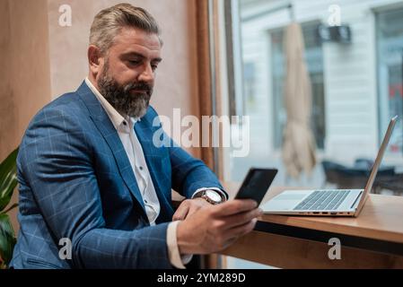 A handsome mature business man using his phone while working on a laptop in a cafe restaurant or office near the window.. Stock Photo