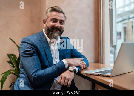 Portrait of a happy old business man sitting at his desk in the office or restaurant cafe near the window and working on a laptop. Stock Photo