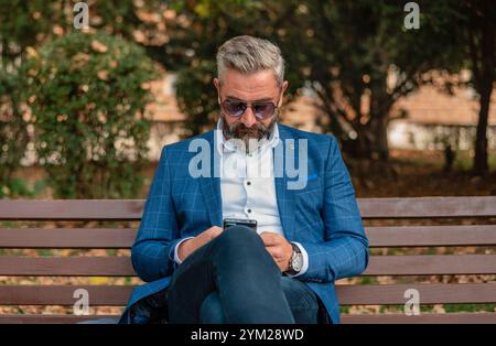A hipster mature business man using mobile phone app texting while sitting on the park bench.  Stock Photo
