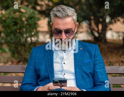 A hipster mature business man using mobile phone app texting while sitting on the park bench.  Stock Photo