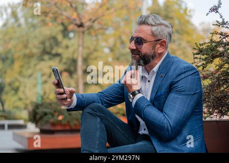 A handsome stylish mature business man making a video call from his mobile phone while sitting on the park bench.  Stock Photo