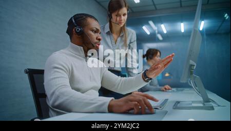 Modern call center office: Two multiethnic technical support specialists wearing headsets talk, discuss work monitoring data on computer. Team of hotline operators working in online customer support. Stock Photo