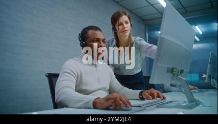 Modern call center office: Two multiethnic technical support specialists wearing headsets talk, discuss work monitoring data on computer. Team of hotline operators working in online customer support. Stock Photo