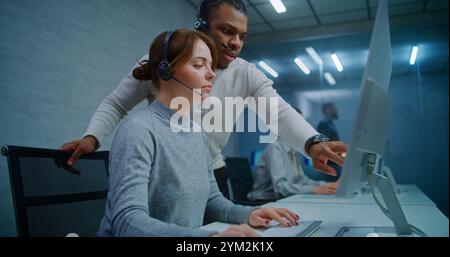 Call center: Female hotline operator in headset monitors data on computer with African American colleague. Multiethnic team of technical support specialists work in online customer support service. Stock Photo