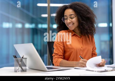 Confident African American businesswoman in modern office, wearing orange shirt, working with laptop, reviewing documents. Represents professionalism, ambition, success in corporate world. Stock Photo