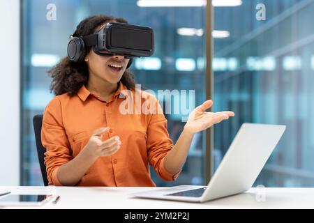 Young African American businesswoman using virtual reality headset while interacting with laptop in modern office. Emphasis on innovation, technology, and professional experience. Stock Photo