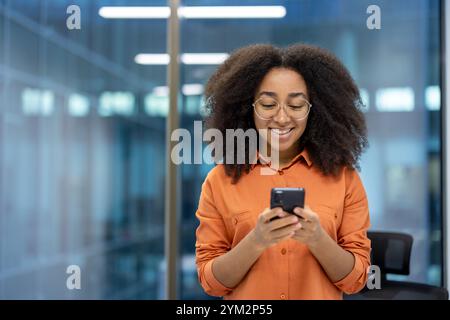 Confident young businesswoman with glasses using smartphone in modern office, smiling at screen during work break. Depicts tech-savvy communication and professional lifestyle. Stock Photo