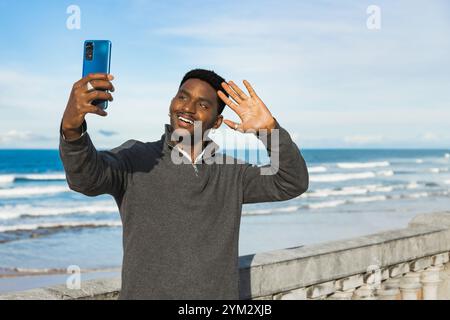 Smiling man waves at the camera while taking a selfie by the ocean. He stands near a stone railing, enjoying the coastal view under a bright sky, capt Stock Photo