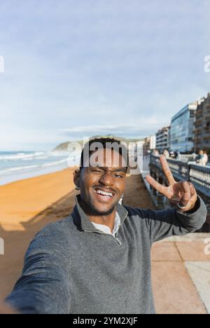 Smiling man winks and holds up a peace sign while taking a selfie along a beachside promenade. The sandy shoreline and urban buildings create a vibran Stock Photo