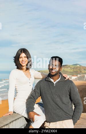 Happy couple poses by the beach on a sunny day, with the ocean waves and a scenic landscape in the background. They stand close, enjoying each other s Stock Photo
