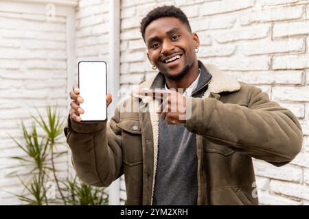Young man in a warm jacket holds up a smartphone with a blank screen, smiling and pointing at it. Great for showcasing apps, digital services, or tech Stock Photo