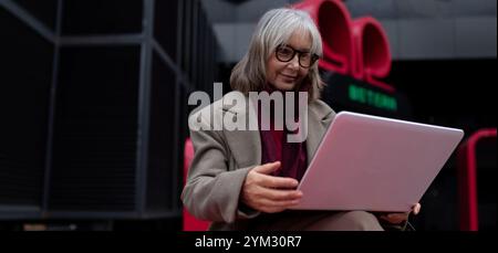Senior businesswoman working on a laptop outside in an urban environment during daytime Stock Photo
