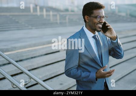 Smiling businessman wearing a suit and glasses talking on phone in front of stairs while holding coffee cup in other hand. Capturing professional moment in urban setting Stock Photo