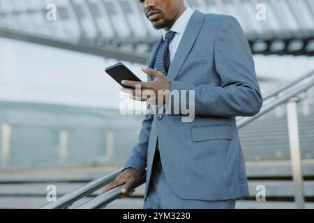 Professional African American man in suit holding smartphone while standing on staircase in modern building. Background includes architectural details of the structure Stock Photo