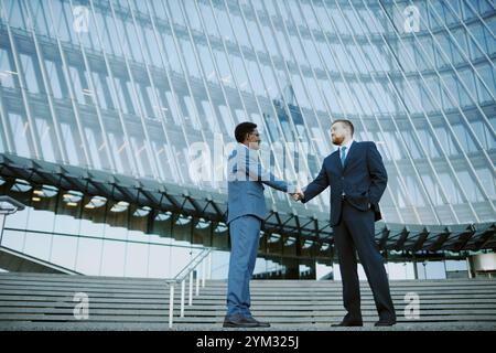 Two businessmen shaking hands while standing outside a modern office building, signifying a professional agreement or partnership Stock Photo
