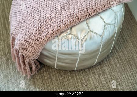 A silver ottoman sits on a carpeted floor with a pink blanket draped over it. The blanket is knitted and has a fuzzy texture. The ottoman is a decorat Stock Photo