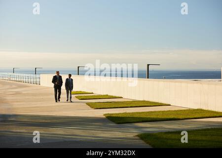 Two business professionals walking side by side on a walkway beside water on sunny day, engaged in conversation. Clear sky and well-maintained grassy area in background Stock Photo