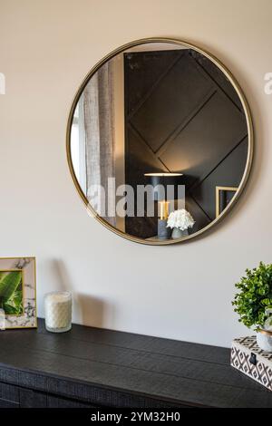 A large round mirror hangs on a wall above a dresser. The dresser is made of dark wood and has a black top. A small table is next to the dresser, and Stock Photo