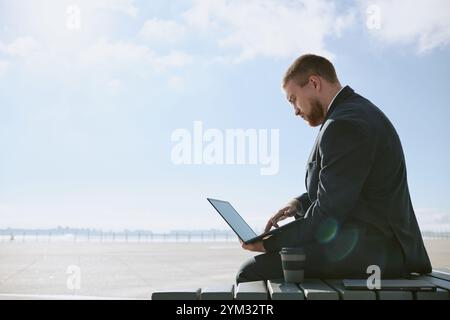 Businessman dressed in suit working on laptop while sitting at airport terminal. Clear sky in background, with coffee cup placed beside him Stock Photo