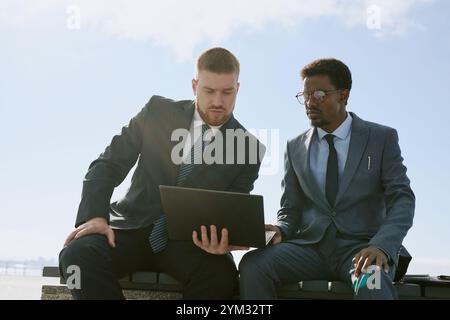 Two businessmen having collaboration while using laptop outdoors during sunny day, sharing discussion. Focus is on interaction between colleagues in professional attire Stock Photo