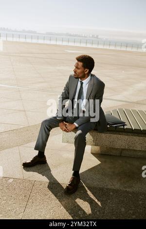 Businessperson in gray suit sitting on bench with ocean background Staring into distance and holding hands together Man with neatly groomed facial hair Stock Photo