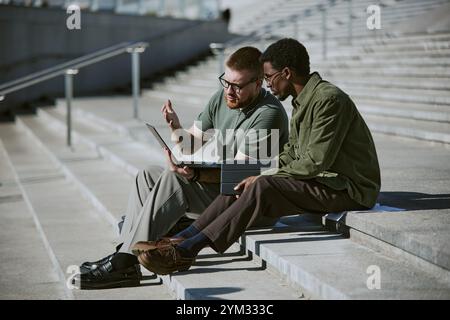Two young men engaged in talking while working on laptop outdoors, seated on steps with serious expressions, focused on task at hand Stock Photo