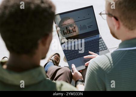 Two men reviewing business reports on laptop screen while sitting outdoors. One man is pointing at screen, showing data and charts, collaborating Stock Photo