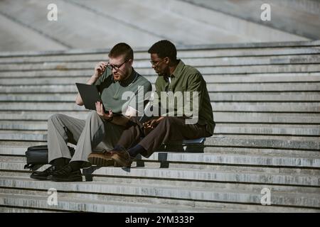 Two men sit on steps, working on laptop and holding notebook. Both engaging in serious discussion, using outdoor environment as workspace Stock Photo