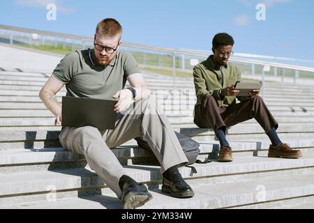 Two men using laptops outdoors while sitting on stairs. One man is writing in notebook while other is typing on his laptop, dressed casually and engaging in work or study Stock Photo