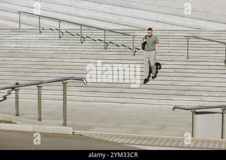 Businessman in green shirt and trousers walking down wide outdoor stairs carrying bag in hand. Clear day with sunlight illuminating stairs and railings in background Stock Photo