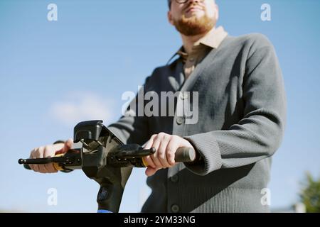 Young man riding electric scooter outside during sunny day wearing a grey cardigan against clear blue sky. Captured in urban setting with relaxed and casual vibe Stock Photo