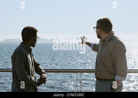 Two professionals standing by railings, gesturing and conversing near urban waterfront with cityscape visible in background, wearing glasses and business attire Stock Photo