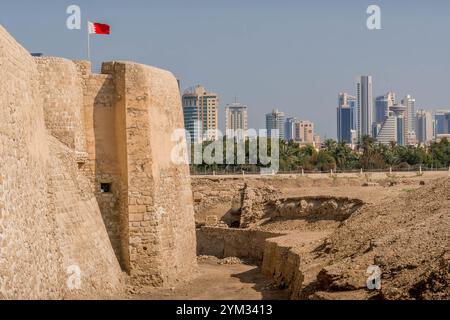 The wall of Qal'at al-Bahrain (Bahrain Fort), an archeological site in Manama, UNESCO World Heritage Site with the city skyline and Bahraini flag. Stock Photo