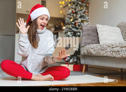 Surprised expression woman in Santa hat having a video call during Christmas Eve, using a laptop at home and waving at the screen. Stock Photo